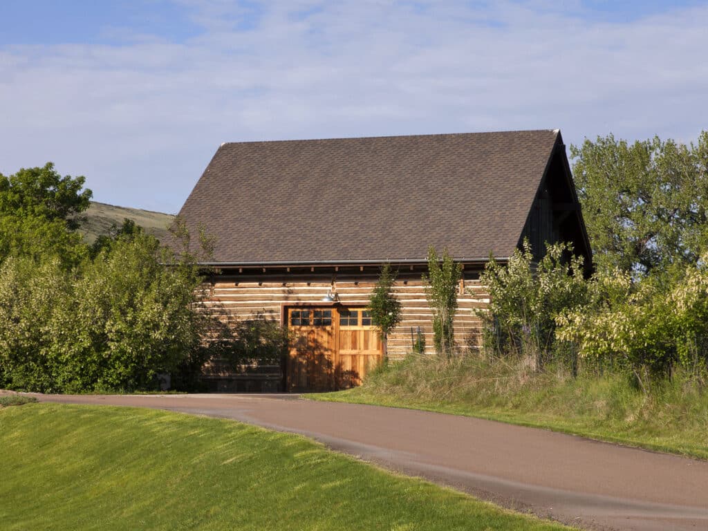 A rustic wooden barn with a pitched roof stands beside a paved road, surrounded by green trees and grass under a partly cloudy sky—ideal recreational land or hunting property.