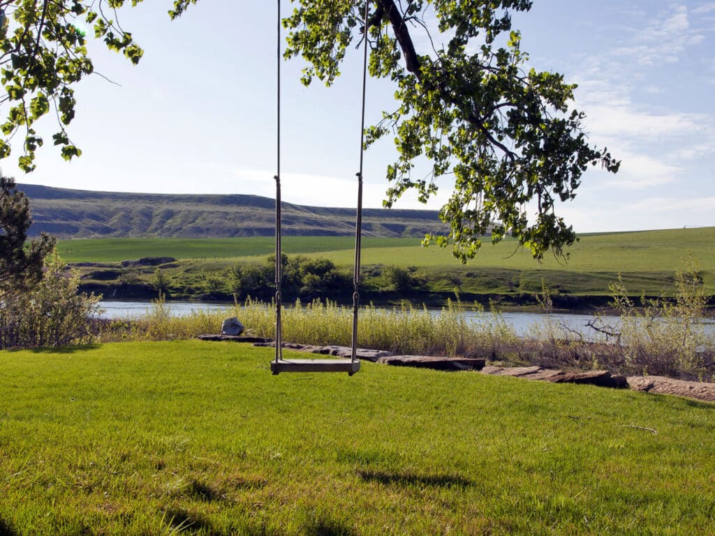 A wooden swing hangs from a tree overlooking a grassy yard on a beautiful hunting property, with a river and green hills in the background under a clear blue sky.