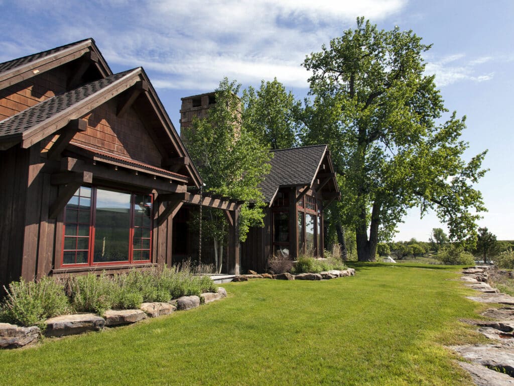 A rustic wooden house with large windows sits on a well-manicured green lawn, surrounded by trees and landscaping stones under a partly cloudy blue sky—an ideal setting for a hunting property or cattle ranch.