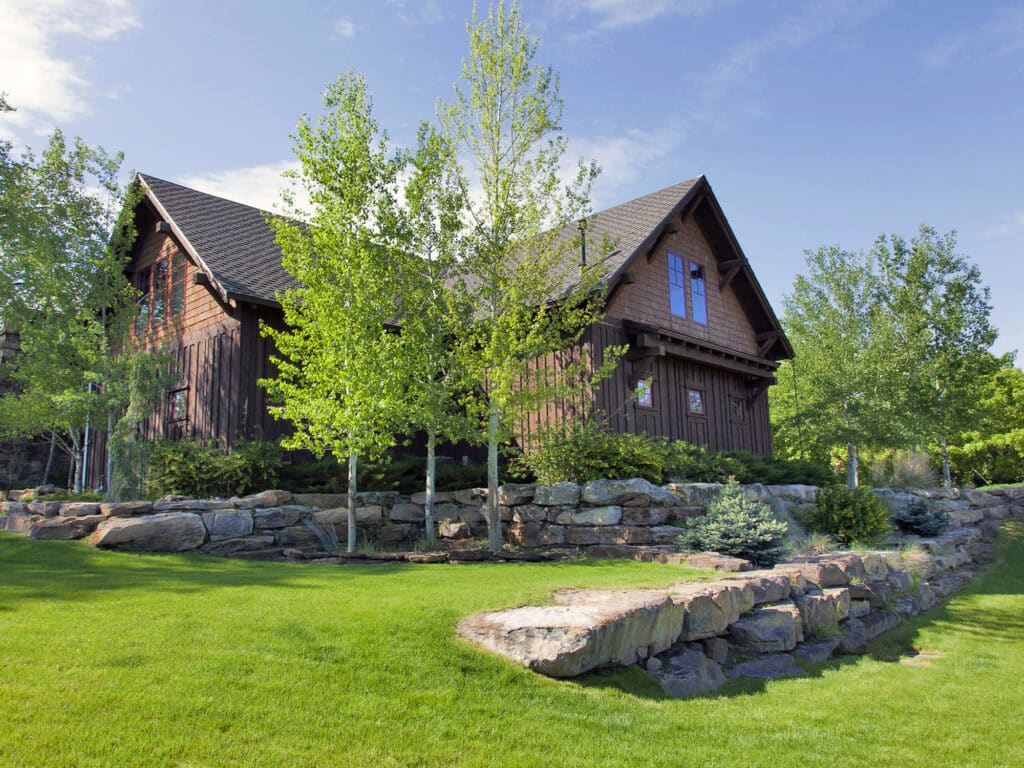 A rustic brown wooden house sits atop a small hill on recreational land, surrounded by green grass, young trees, and large stone landscaping under a clear blue sky.