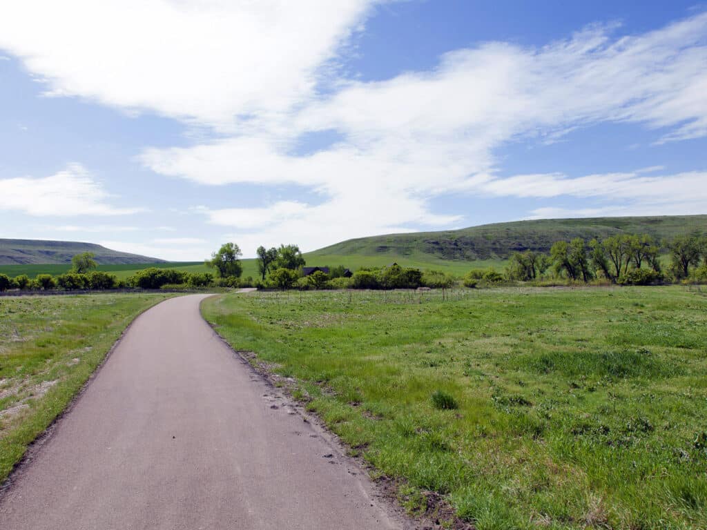 A paved path curves through a green grassy field with scattered trees, winding through recreational land for sale and leading toward rolling hills under a partly cloudy blue sky.