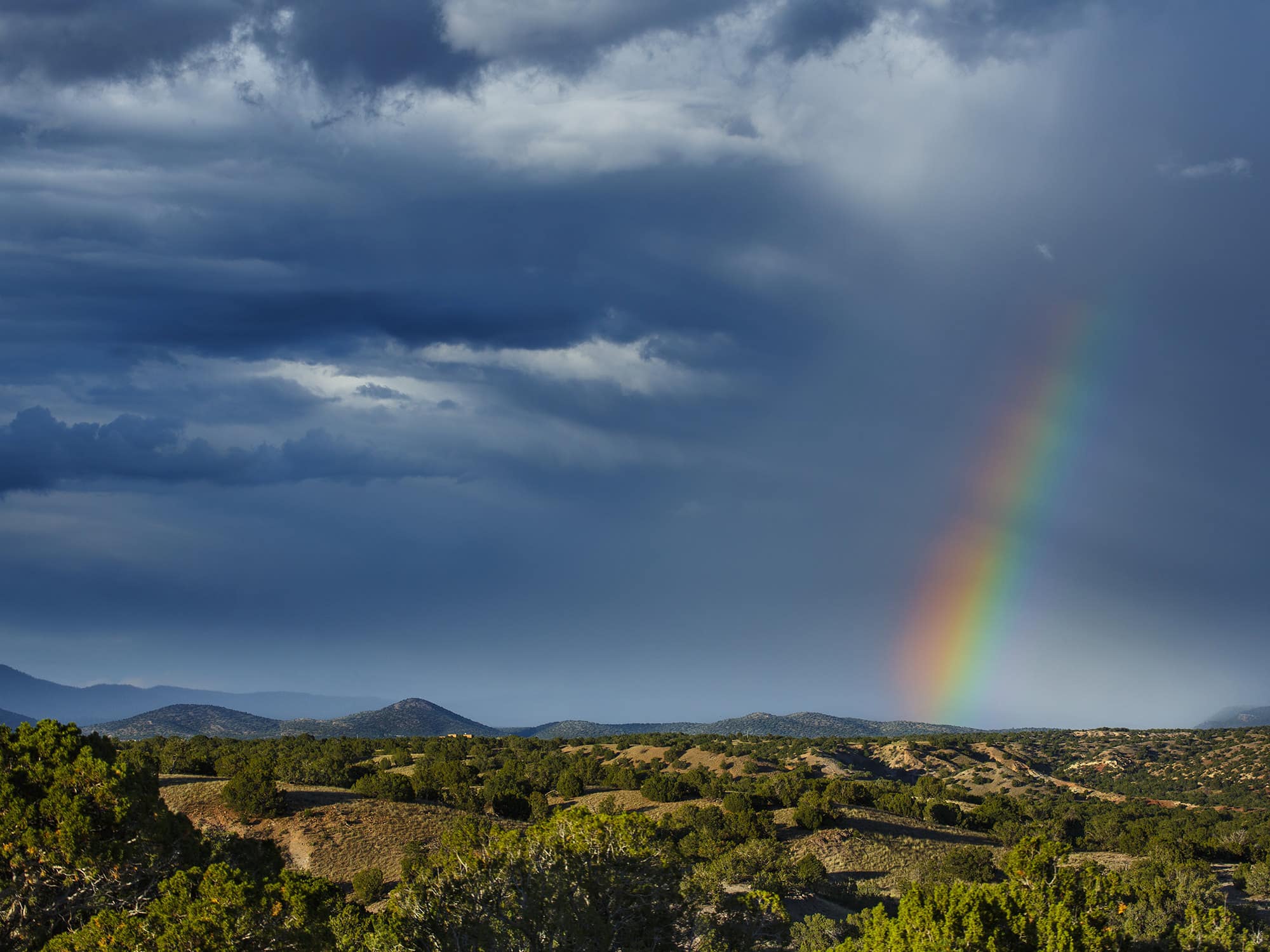 A vibrant rainbow appears against a dark, cloudy sky over a hilly, green landscape with distant mountains—an ideal setting for recreational land or a cattle ranch.