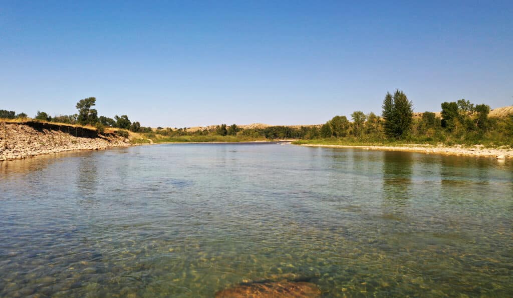 A clear, shallow river flows gently under a bright blue sky, bordered by rocky banks and lush green trees—an ideal recreational land with distant hills visible on the horizon.