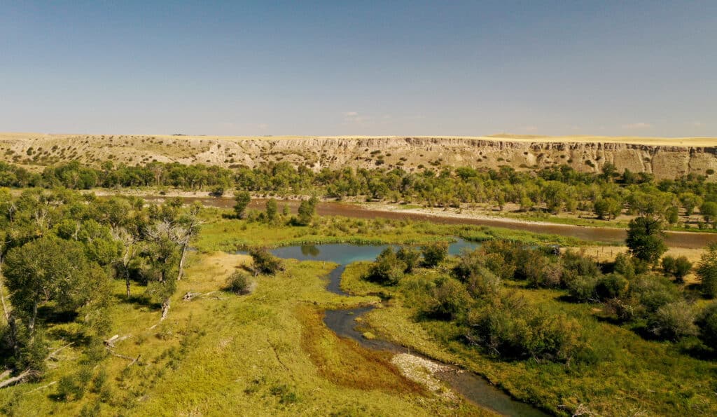 A river winds through a green, tree-filled valley with a rocky, beige plateau in the background under a clear blue sky—an ideal hunting property or recreational land.