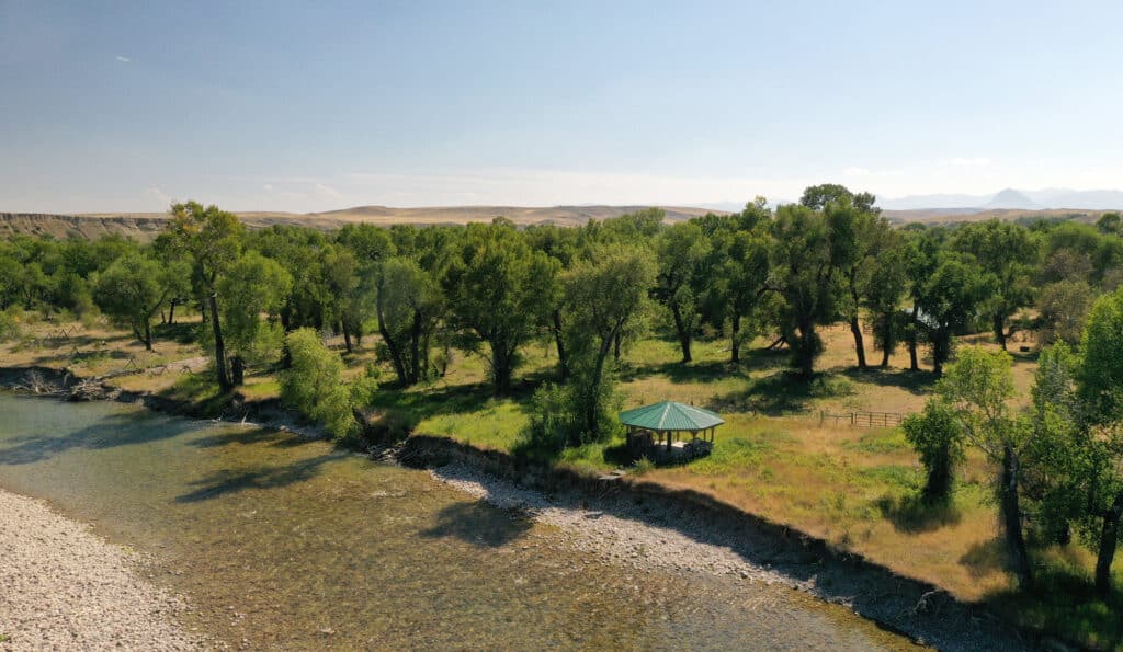 A small gazebo with a green roof sits on recreational land near a rocky riverbank, surrounded by tall trees under a clear blue sky with distant hills in the background.