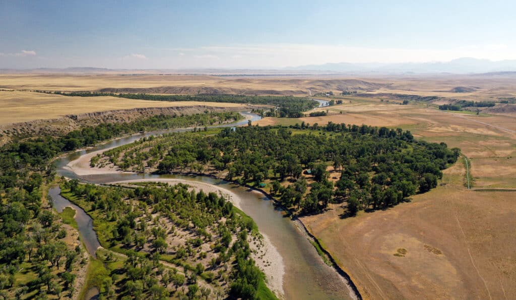 Aerial view of a winding river surrounded by dense green trees, flowing through recreational land and golden fields with distant hills under a clear blue sky—an ideal ranch for sale.