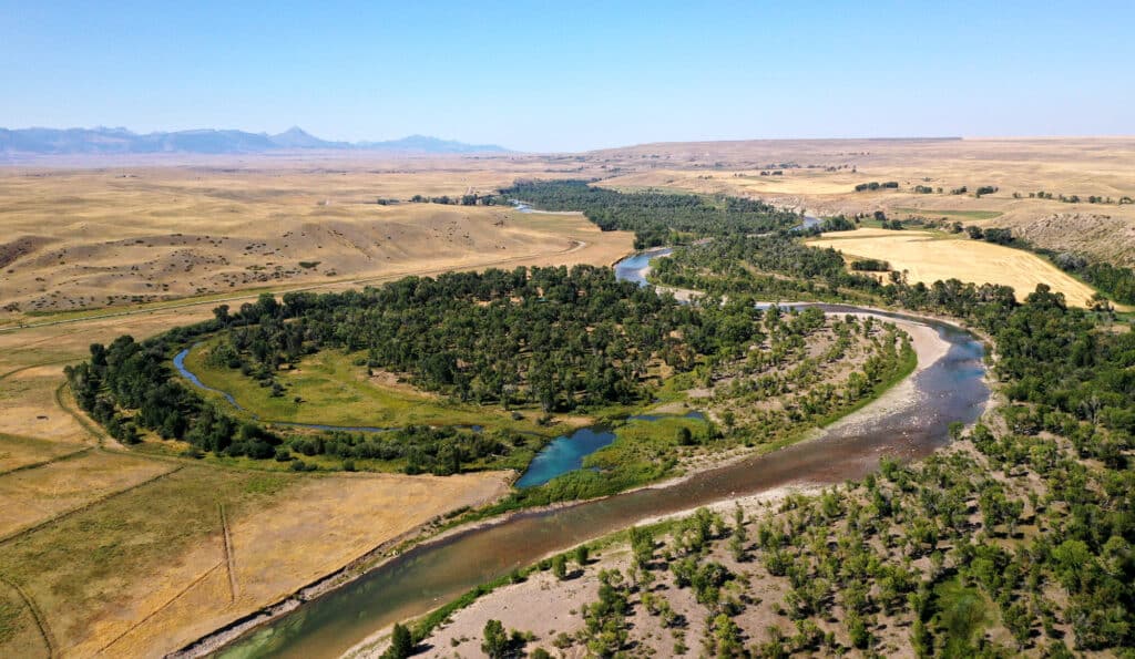 Aerial view of a winding river flowing through a green, tree-lined area on a hunting property, surrounded by dry, golden fields and distant mountains under a clear blue sky.