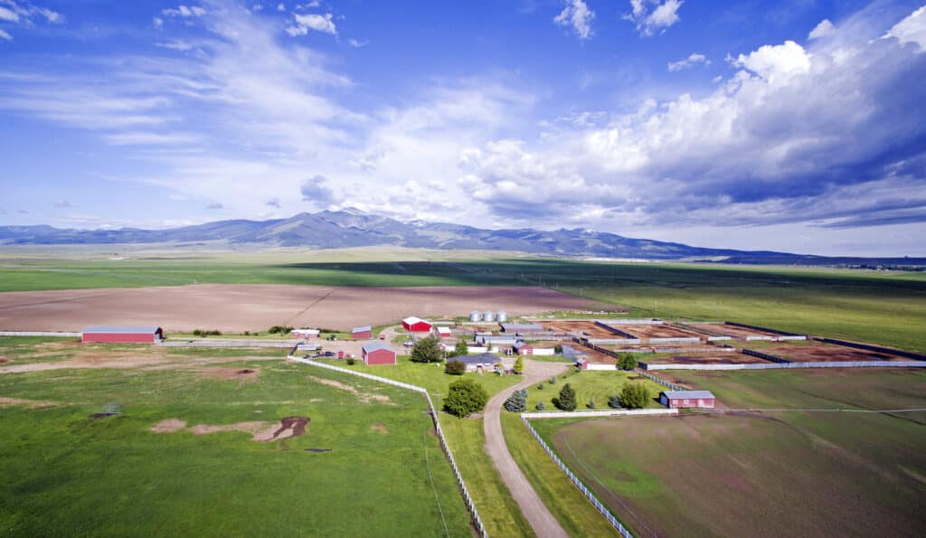 Aerial view of a large farm with barns and fields, surrounded by green and brown farmland—ideal recreational land or hunting property—with mountains in the background beneath a partly cloudy blue sky.