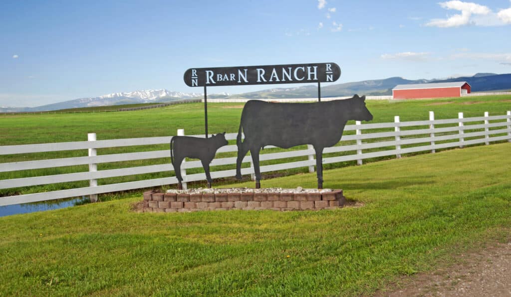 A metal sign with silhouettes of a cow and calf reads R Bar N Ranch, marking this cattle ranch on a grassy lawn by a white fence, with fields, mountains, and a red barn visible in the background.