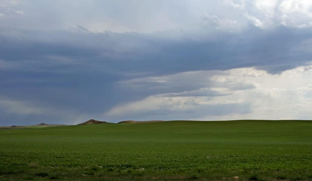 Wide open green field with low hills in the distance under a dramatic sky filled with dark, heavy clouds—perfect recreational land or potential hunting property as a storm approaches.