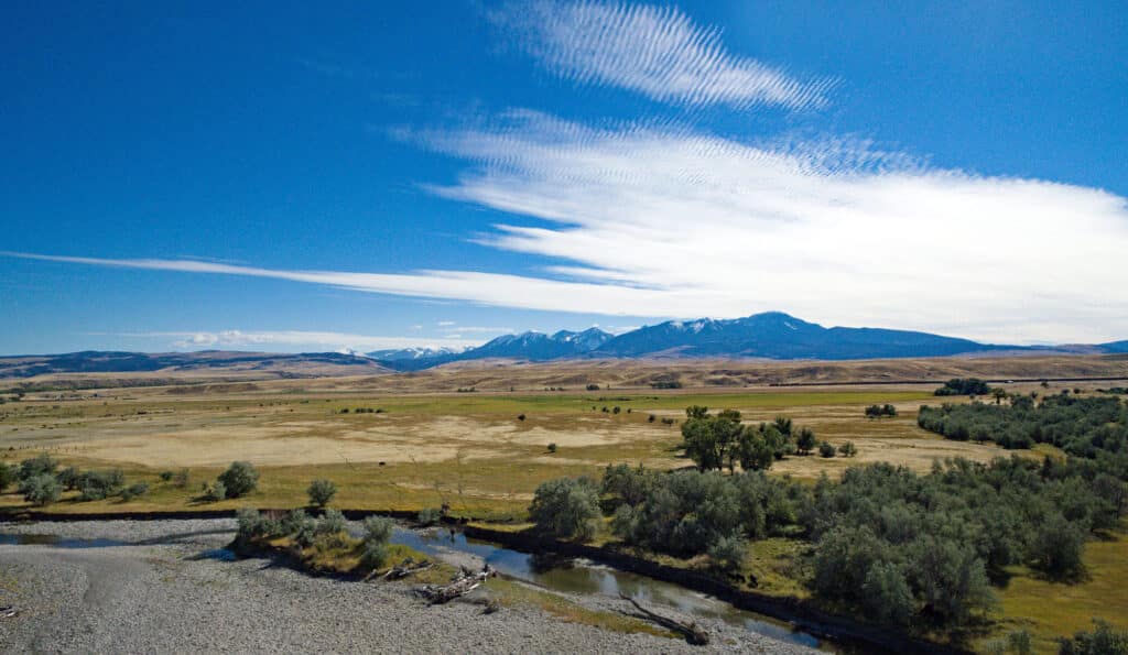 A wide view of recreational land with scattered trees, a small river in the foreground, and distant mountains under a bright blue sky with streaks of white clouds. Perfect hunting property for nature lovers.