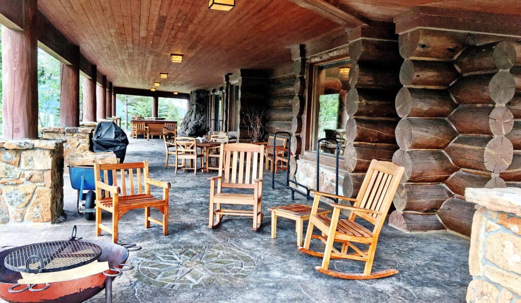 A rustic log cabin porch with wooden rocking chairs, a fire pit, grill, and tables, surrounded by stone pillars and forest scenery—perfect for enjoying a recreational land escape or relaxing at your hunting property.