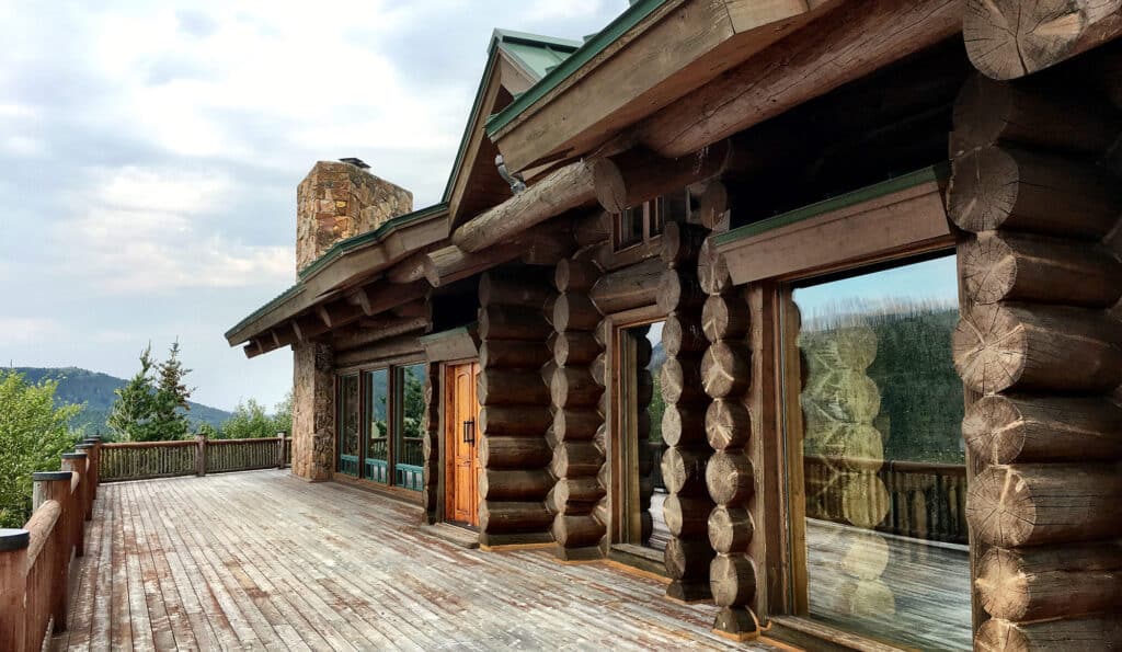 A large wooden log cabin with a spacious, weathered deck, large windows reflecting trees, and a stone chimney, set in a mountainous, forested area under a cloudy sky—perfect for use as a hunting property or recreational land.
