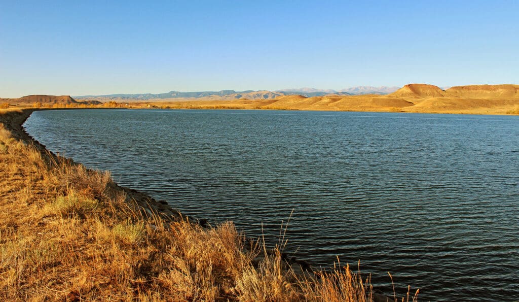 A wide river or lake curves through dry, golden grasslands under a clear blue sky, with distant mountains and brown hills on the horizon—perfect recreational land or cattle ranch opportunity.