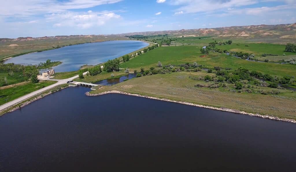 Aerial view of a reservoir with a dam and road, surrounded by green fields, trees, and gently rolling hills—ideal recreational land or hunting property under a blue sky with scattered clouds.