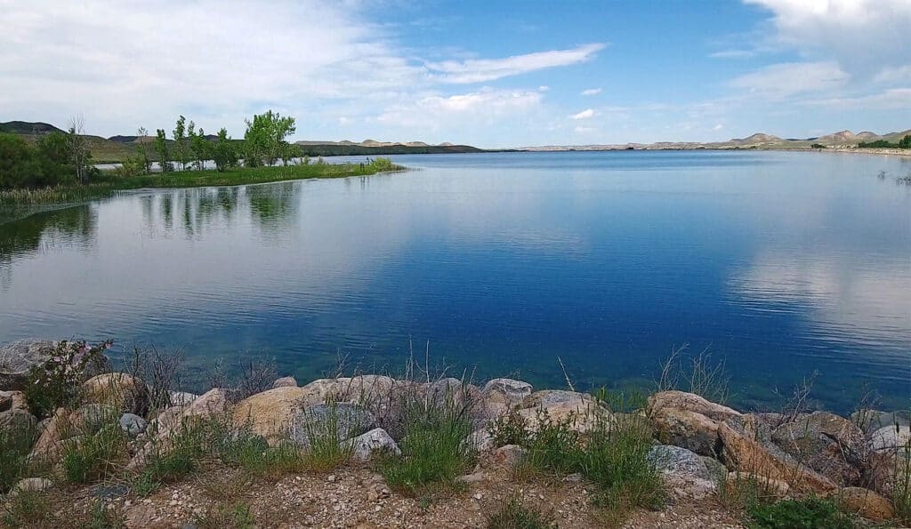 A calm lake with clear blue water, bordered by rocks and grass in the foreground, with a few green trees on the left and distant hills under a partly cloudy sky—ideal for recreational land or a serene hunting property.