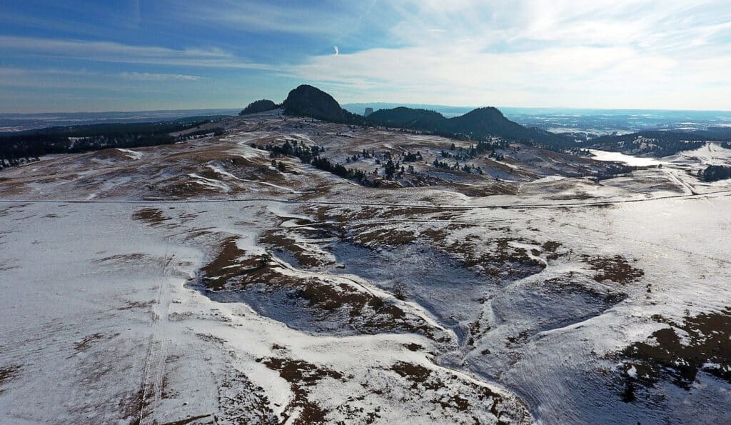 A wide landscape with snow-dusted hills and patches of brown grass leads to rocky formations and distant mountains under a mostly clear sky—ideal hunting property or ranch for sale.