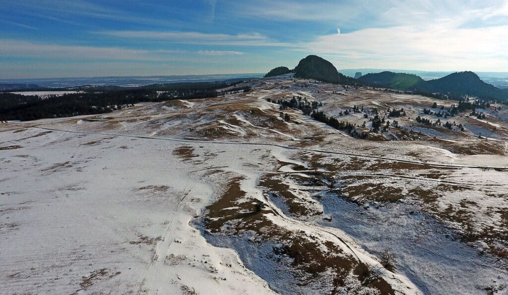 Aerial view of a snow-dusted landscape with rolling hills and scattered trees, leading to a large, rocky outcrop under a blue sky—an ideal cattle ranch or hunting property.