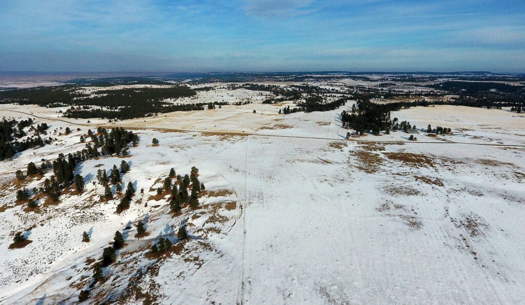 A wide aerial view of a snow-covered cattle ranch with rolling hills, scattered trees, and patches of brown grass under a partly cloudy blue sky. A narrow road cuts through this picturesque land for sale.