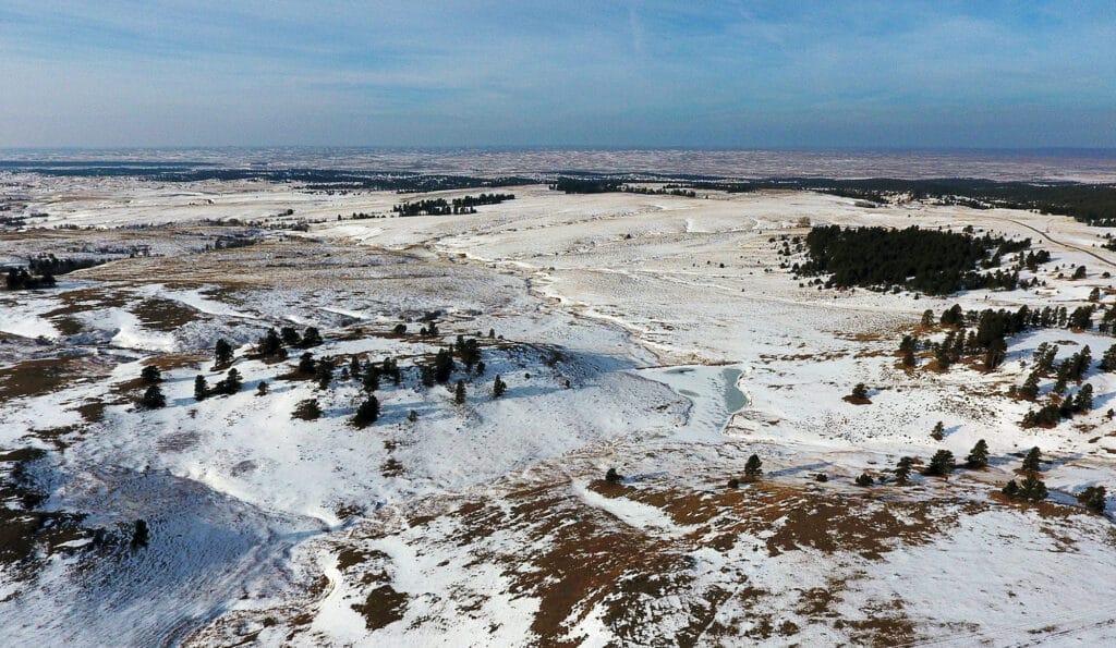 Aerial view of a vast, snow-covered landscape with patches of exposed ground, scattered trees, and distant hills under a blue sky—ideal land for sale or a picturesque cattle ranch.