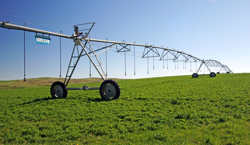 A large center pivot irrigation system stands in a green field under a clear blue sky, with rolling hills visible in the background—ideal features for those seeking ranch for sale or hunting property.