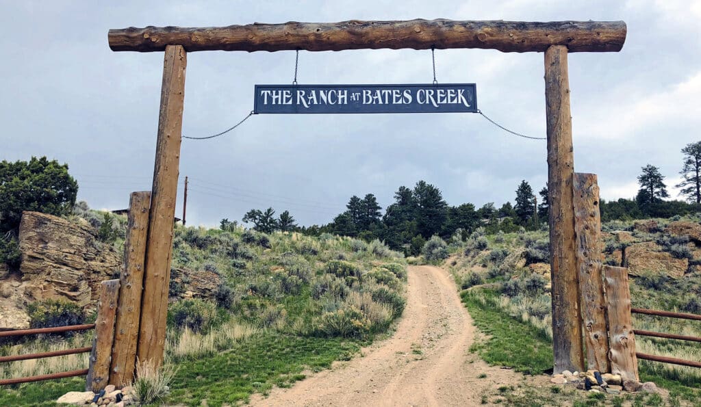 A rustic wooden entrance gate with a sign reading The Ranch at Bates Creek stands over a dirt road, welcoming you to this scenic cattle ranch nestled in a grassy, tree-dotted landscape beneath a cloudy sky.