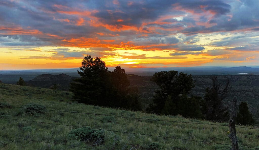 A vibrant sunset with orange and yellow hues illuminates a cloudy sky above distant mountains, silhouetted trees, and a grassy field with a wire fence—ideal scenery for a recreational land or picturesque cattle ranch.