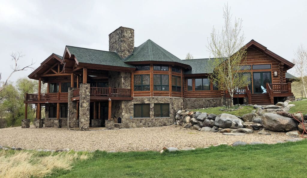 A large rustic log cabin with stone accents, multiple balconies, large windows, and a green roof sits on a gravel driveway surrounded by rocks, lawn, and sparse trees—ideal as a hunting property or unique land for sale.