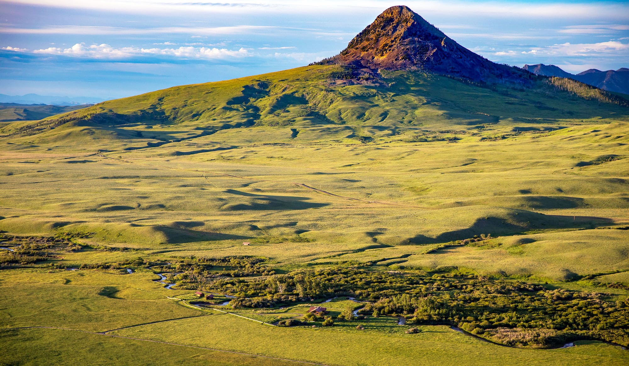 Green grassy plains with a solitary, conical hill rising in the background under a blue sky, sunlight casting shadows over this scenic recreational land for sale. Small clusters of trees and a winding stream enhance the picturesque foreground.