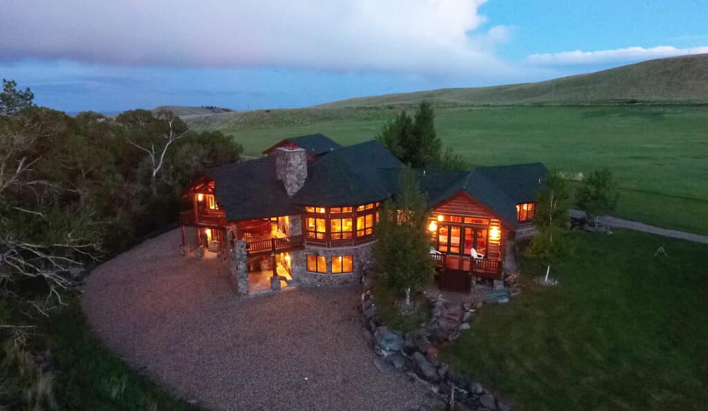 A large, rustic house with glowing windows sits surrounded by trees, grass, and hills at dusk on a peaceful hunting property, creating a warm and inviting atmosphere in the rural landscape.
