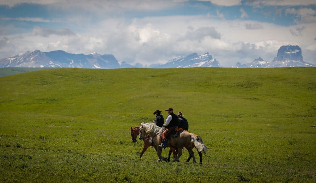 Three people wearing cowboy hats ride horses across a vast green field on a cattle ranch, with rugged mountains rising in the distance under a partly cloudy sky.