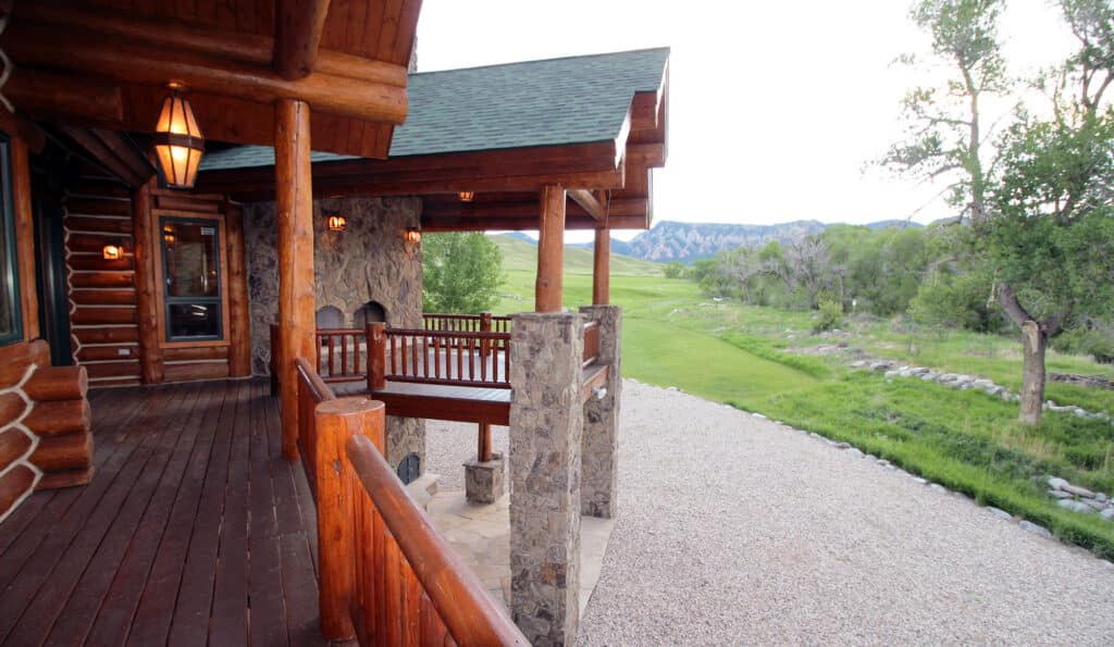 A wooden log cabin porch with stone pillars overlooks a gravel driveway, green lawn, trees, and distant mountains under a bright, clear sky—perfect for those seeking a hunting property or ranch for sale.