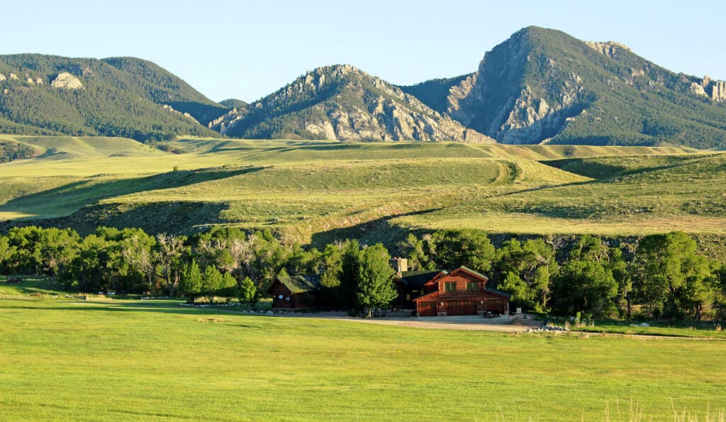 A rustic wooden lodge surrounded by trees sits in a wide, green valley with rolling hills—perfect as a hunting property or cattle ranch—with dramatic, rocky mountains in the background under a clear blue sky.