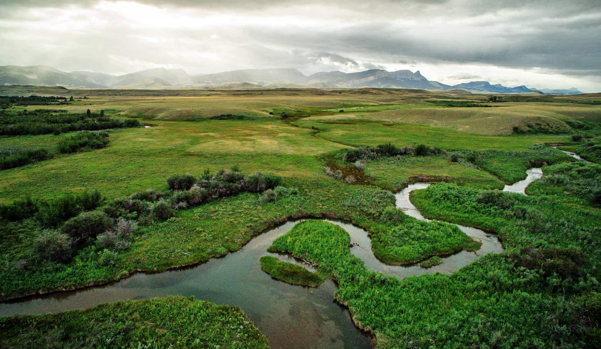 A winding river flows through lush green fields and shrubs under a cloudy sky, with distant mountains on the horizon. This vast, serene landscape is ideal recreational land or hunting property, perfect for those seeking land for sale.