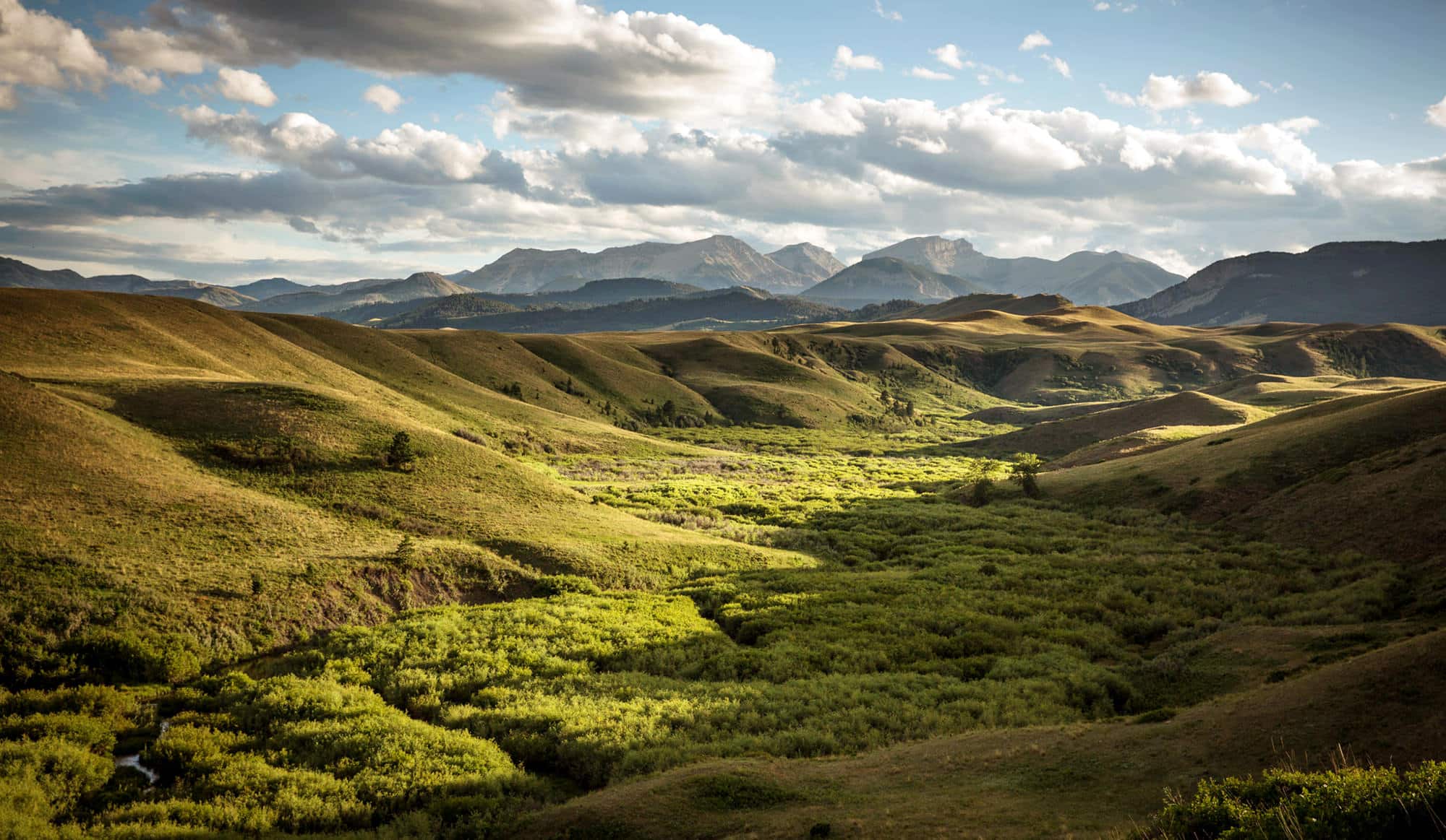 Sunlit rolling green hills and a lush valley stretch toward distant, rugged mountains under a partly cloudy sky. Dramatic contrasts of shadow and light highlight the recreational land, perfect for those seeking hunting property in a scenic landscape.