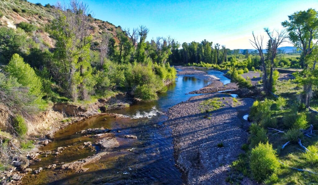A clear river winds through a forested landscape with lush green trees on both banks, a rocky shoreline, and hills in the background under a bright blue sky—perfect as a scenic hunting property or cattle ranch for sale.
