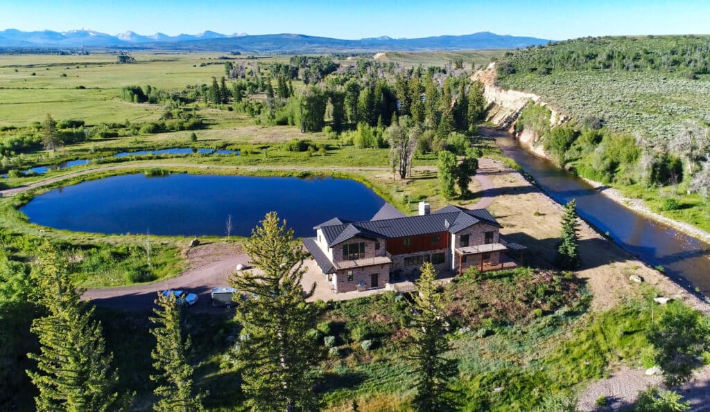 A large ranch for sale sits beside a pond and river, surrounded by green fields, trees, and distant mountains under a clear blue sky.