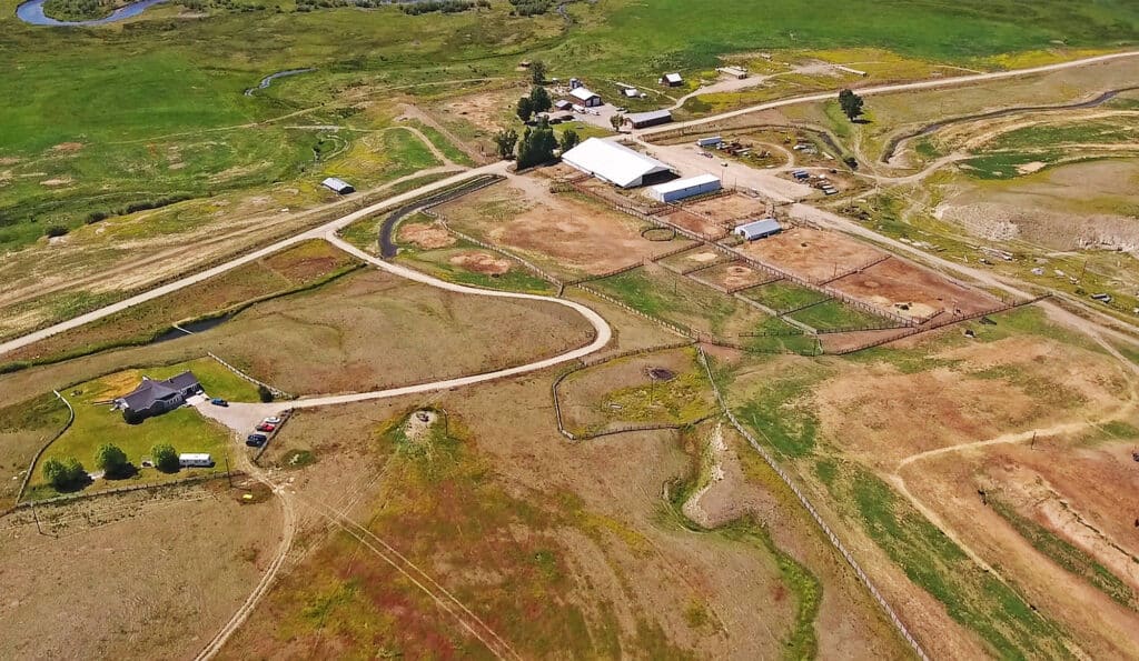 Aerial view of a rural ranch for sale with barns, fenced areas, dirt roads, scattered vehicles, and a house surrounded by green fields and patches of dry land.