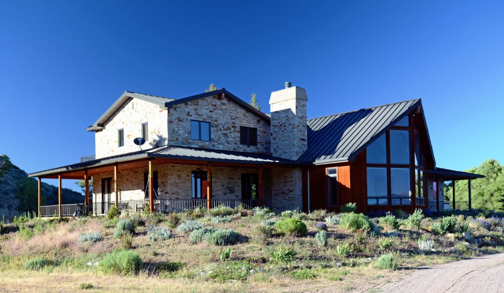 A modern two-story house with stone and wood exterior, large windows, a black metal roof, and a wraparound porch sits on a grassy plot of recreational land under a clear blue sky.