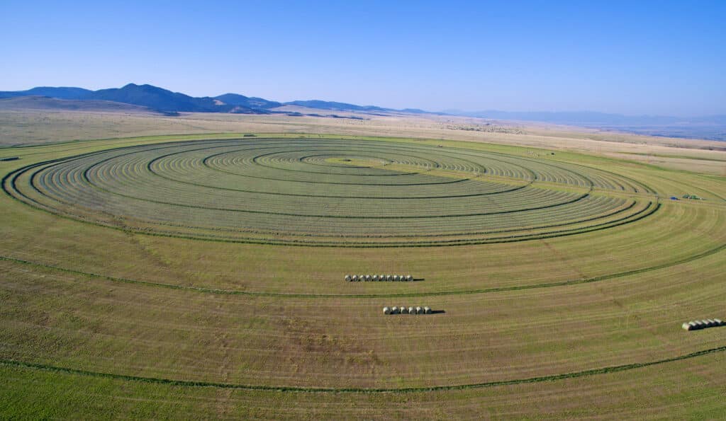 Aerial view of a large, circular crop field with concentric green rings, ideal recreational land surrounded by flat farmland and distant mountains under a clear blue sky. Several hay bales are lined up near the center of the field.