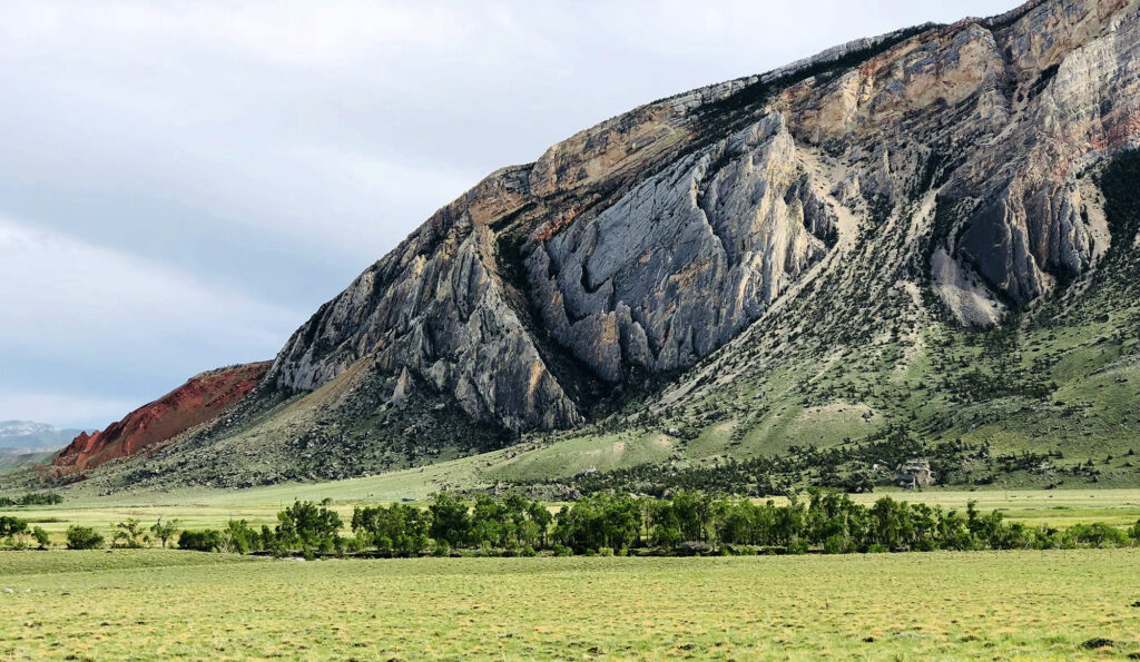 A rugged, multicolored mountain with steep, jagged rock faces rises above a green valley with scattered trees and grass—an ideal backdrop for hunting property or cattle ranch land under a cloudy sky.