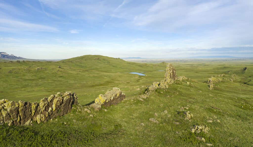A scenic view of rolling green hills with rocky outcrops in the foreground and a small blue pond in the distance under a blue sky with scattered clouds, perfect for a cattle ranch or those seeking land for sale.