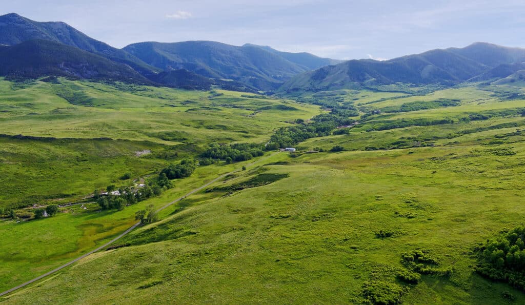 Aerial view of a lush green valley with scattered trees and small buildings, ideal as a cattle ranch or hunting property, surrounded by rolling hills and distant mountains under a clear blue sky.
