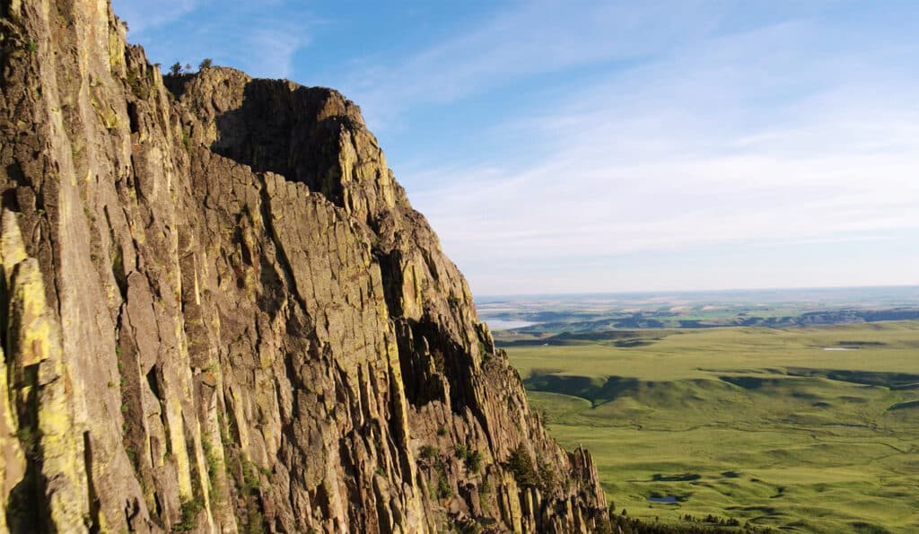 A steep rocky cliff rises on the left, overlooking a vast green plain ideal for a cattle ranch that stretches into the distance under a blue sky with wispy clouds.