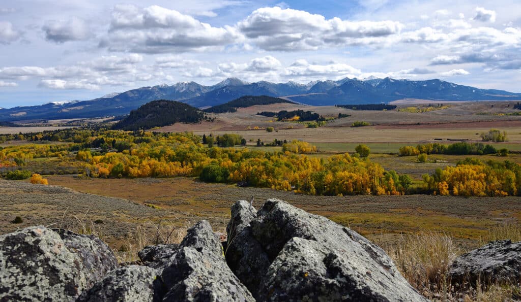 Rocky foreground overlooking a wide valley with scattered yellow and green trees, open fields ideal for a cattle ranch, and a distant range of snow-capped mountains under a partly cloudy sky.