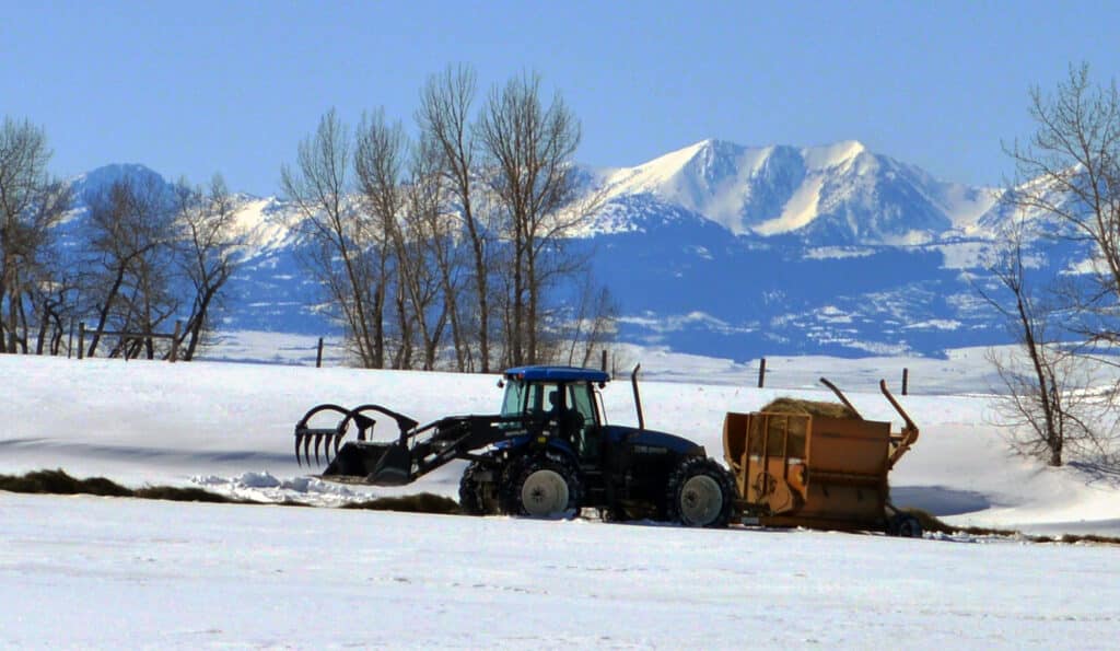 A tractor with a front loader attachment works on recreational land in a snow-covered field, with bare trees and snow-capped mountains in the background under a clear blue sky.