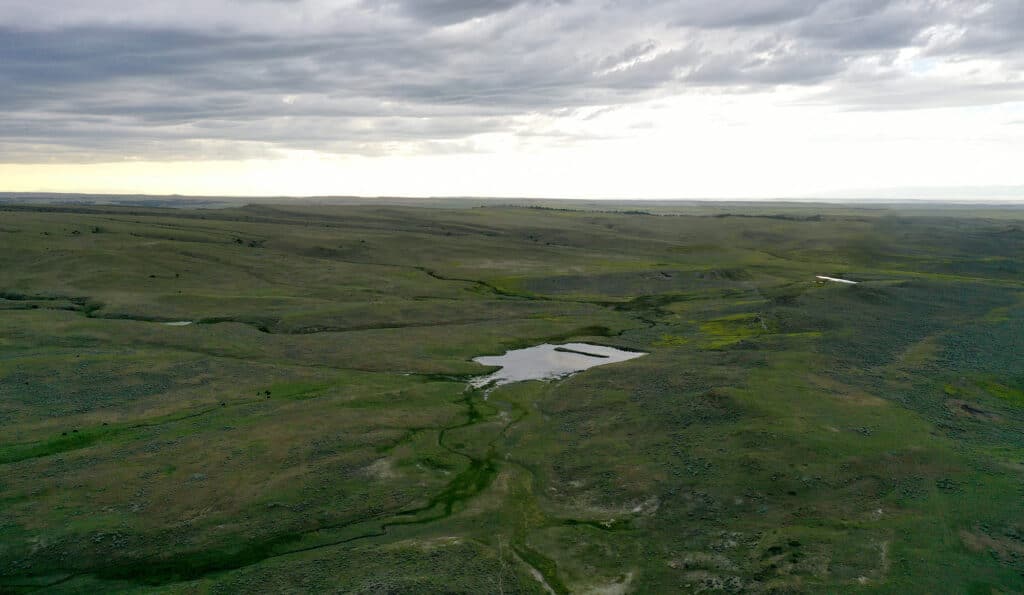 A wide, grassy plain stretches under a cloudy sky at sunset, with a small reflective pond and patches of greenery among rolling hills—ideal recreational land or potential ranch for sale.