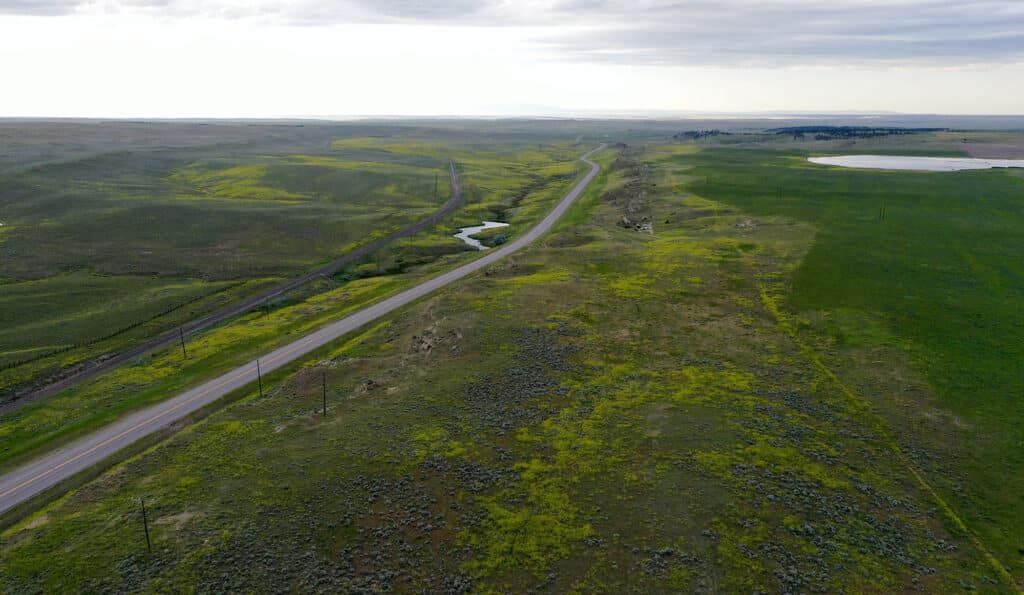Aerial view of a long, empty road winding through vast green fields and rolling hills under a cloudy sky, highlighting recreational land with small patches of water visible in the landscape.