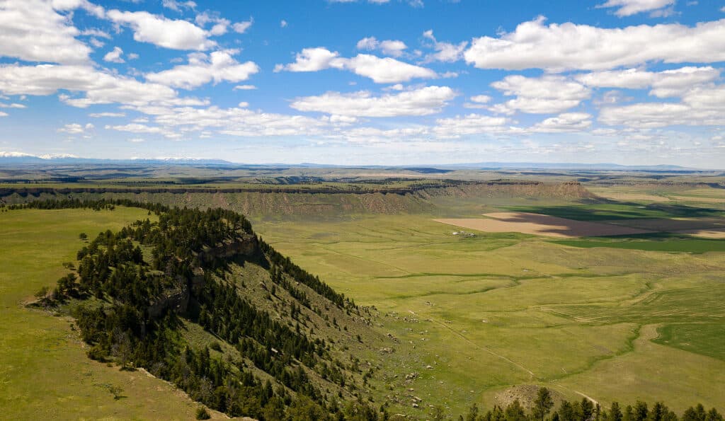 Aerial view of a green plateau with scattered trees, dramatic cliffs, and wide open plains under a blue sky. Perfect as recreational land or hunting property, with fields and distant mountains visible on the horizon.