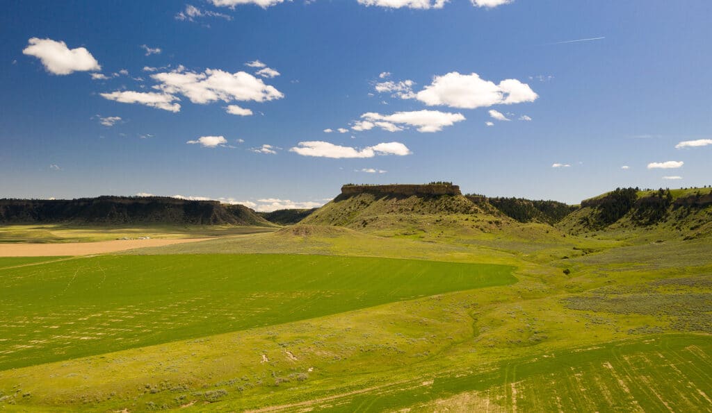 A wide green field stretches toward a flat-topped hill under a bright blue sky with scattered clouds, ideal as recreational land or cattle ranch, surrounded by gentle slopes and distant ridges.